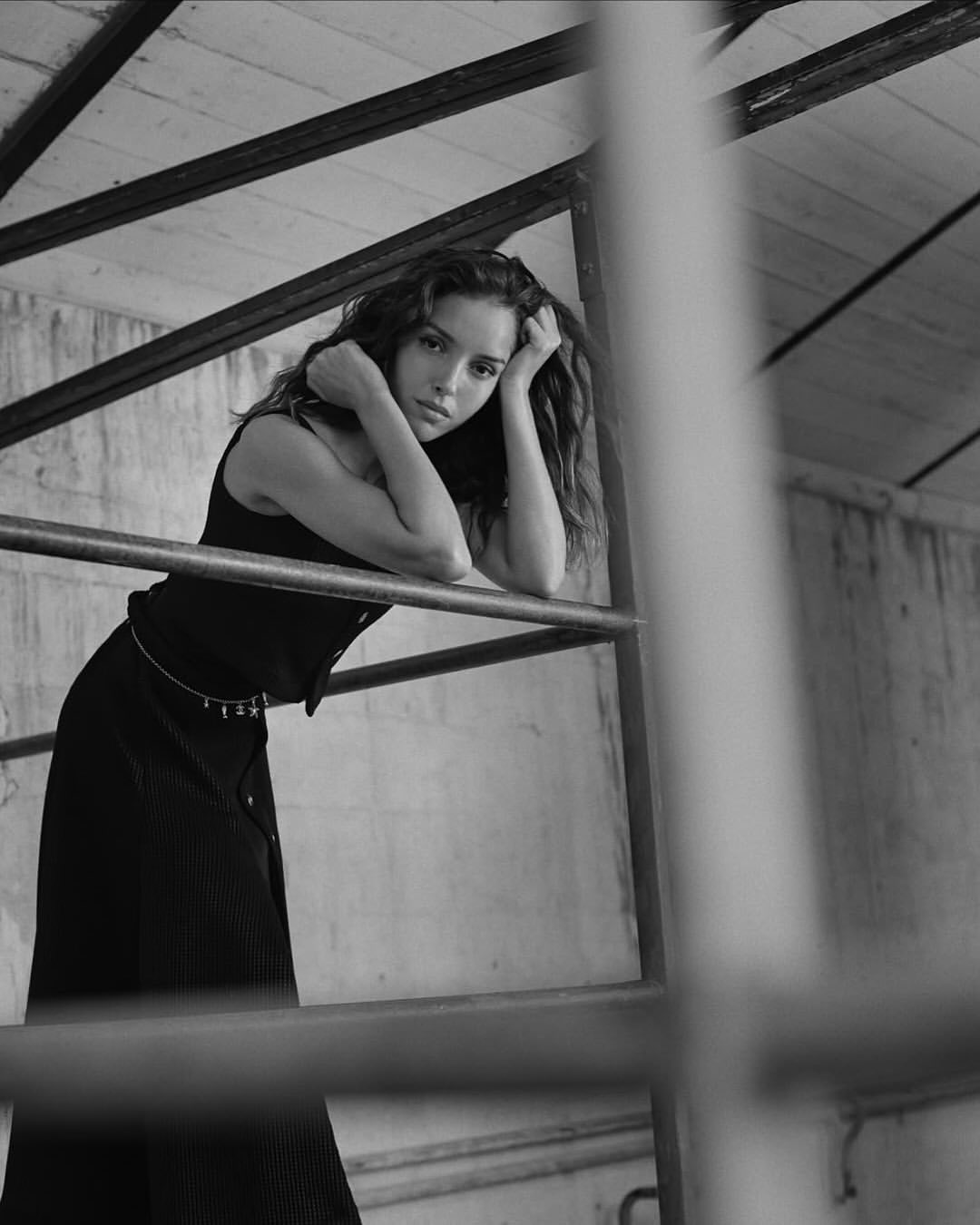 Black and white portrait of a woman with long, wavy hair leaning on a metal structure indoors with a serious facial expression.