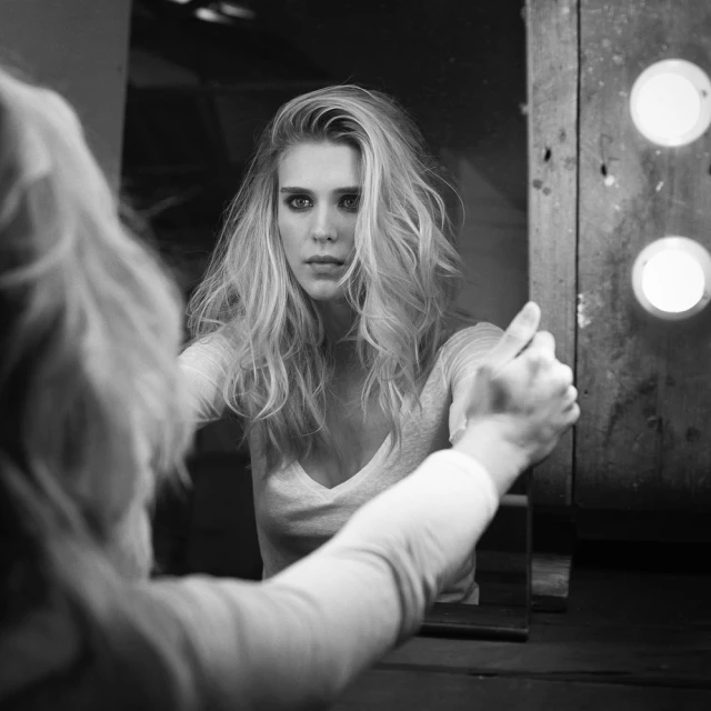 Black and white portrait of an adult female with long blonde wavy hair looking in a vanity mirror with two lights, indoors.