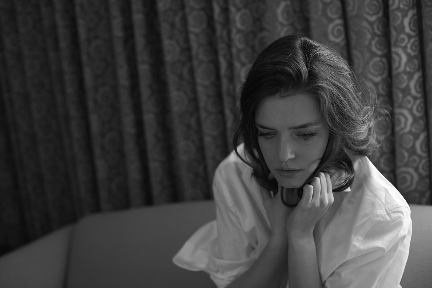 Black and white portrait of a woman in a white shirt sitting indoors with dark patterned curtains behind her.