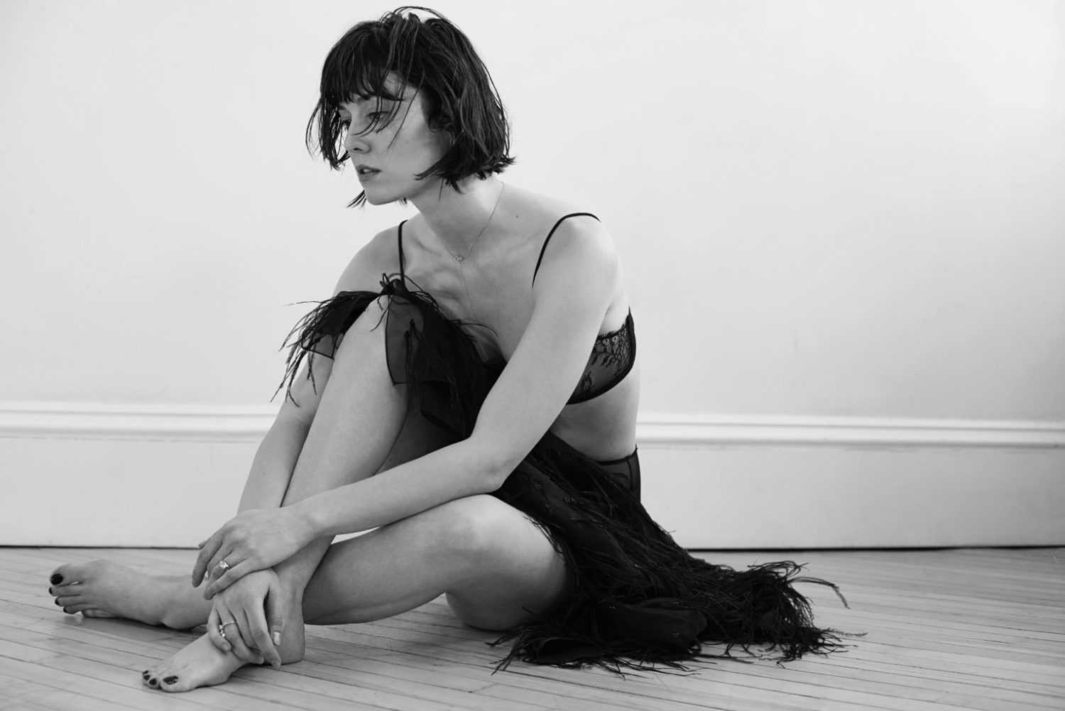 Black and white portrait of a young adult woman with short dark hair in a black lace bra and fringed skirt sitting on the floor.