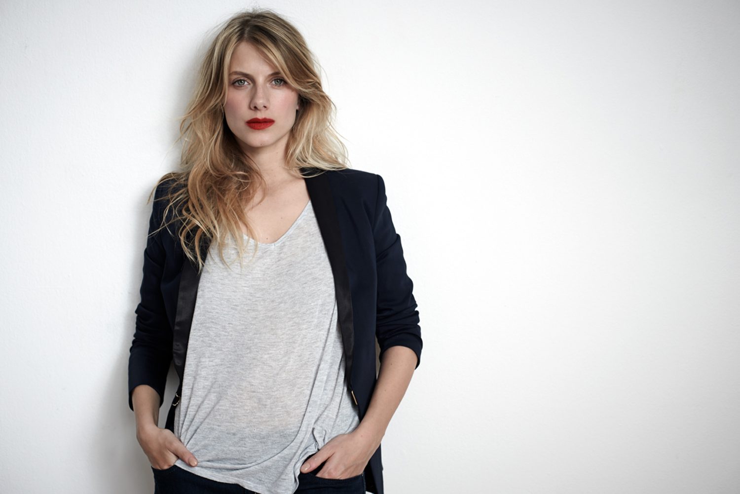 Blonde woman with red lipstick posing against white background in a casual t-shirt and jacket, looking toward the camera.