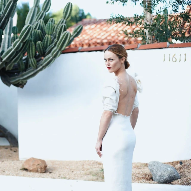 Woman in elegant white dress stands outdoors near cactus and white building, looking over shoulder with bright red lipstick.
