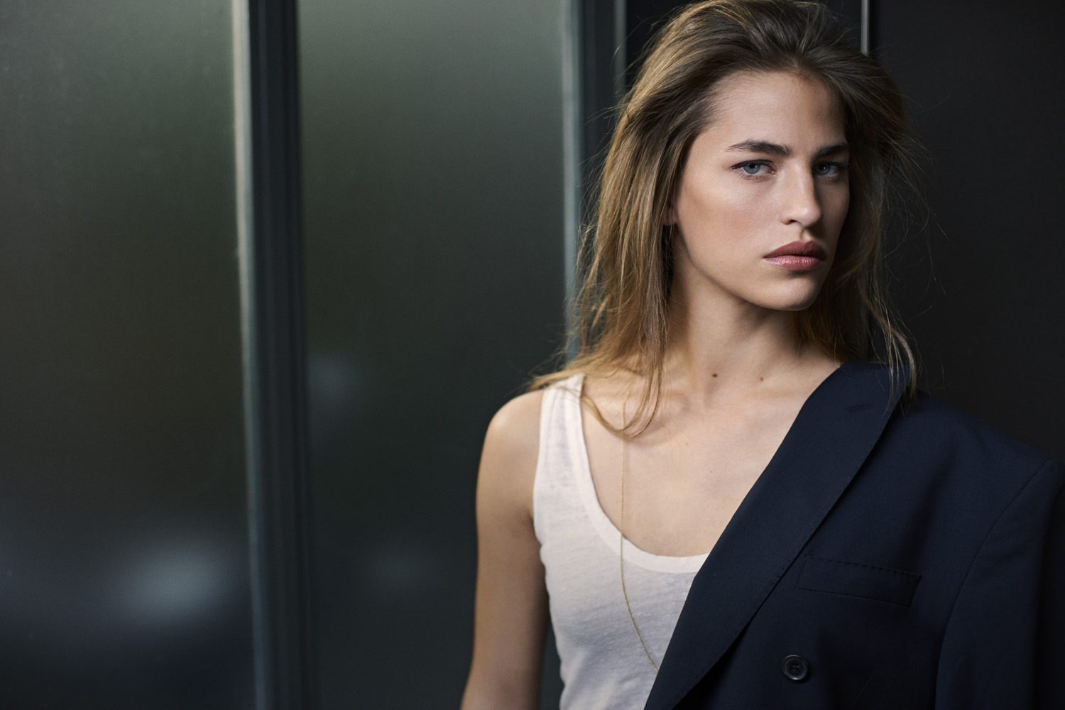 Portrait of a woman wearing a white tank top and a dark blazer draped over one shoulder against a dark wall