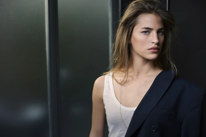 Portrait of a woman wearing a white tank top and a dark blazer draped over one shoulder against a dark wall
