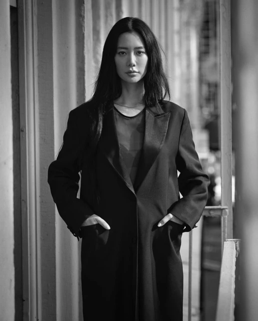Black and white portrait of a stylish adult woman with long dark hair, wearing a long black coat with her hands in her pockets.