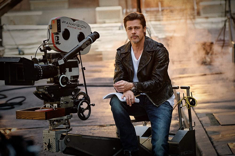 Adult male actor Brad Pitt sits near a Panavision camera on set, wearing a leather jacket and jeans with a script in hand.