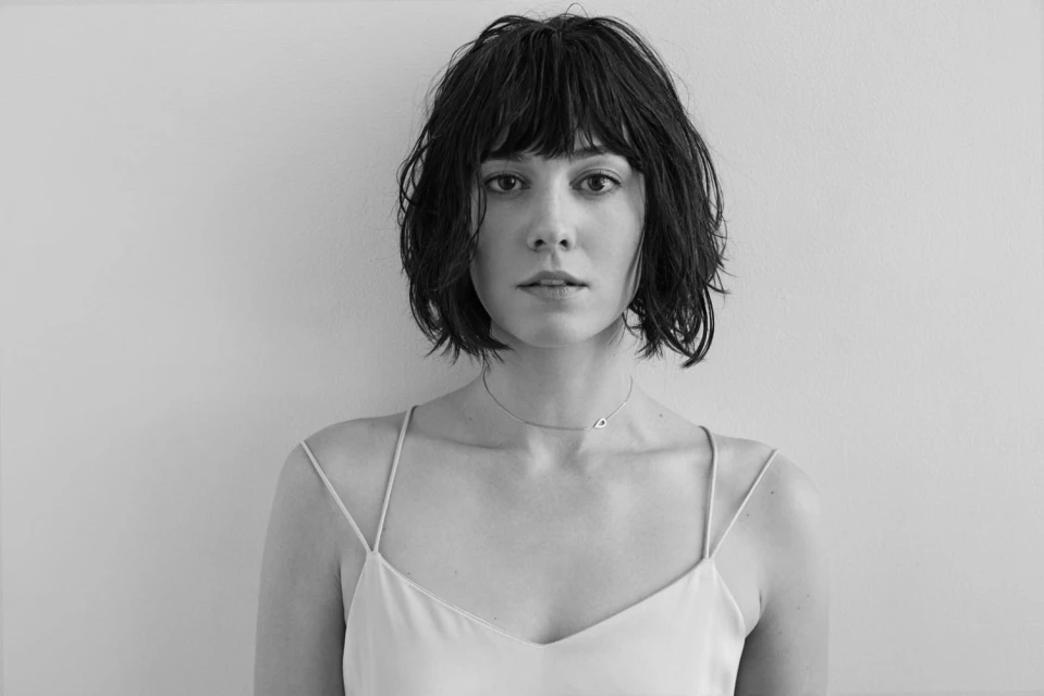 Monochrome portrait of a fair-skinned adult woman with dark, shaggy hair, wearing a light-colored spaghetti-strap top against a plain background.