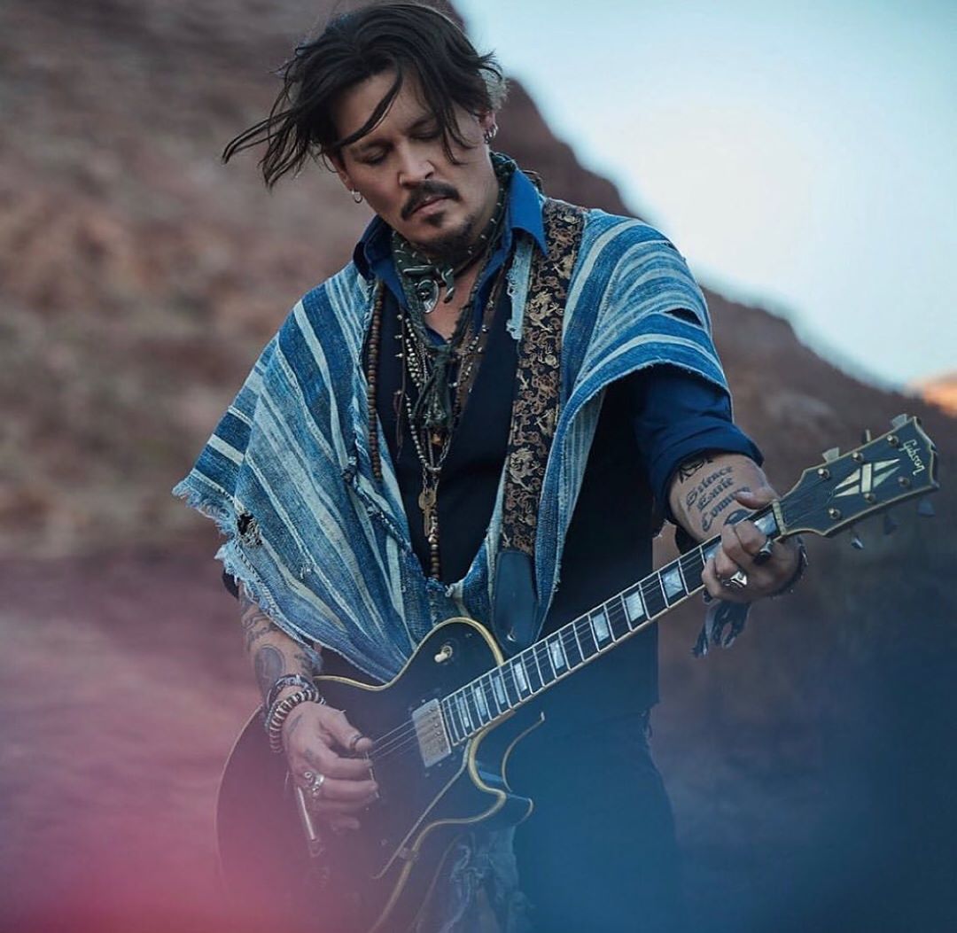 Johnny Depp plays electric guitar in an outdoor, desert-like setting, wearing a shawl, necklaces, and rings, with visible tattoos.