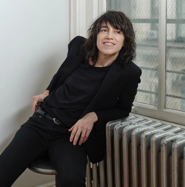 Portrait of a smiling adult woman with dark, wavy hair, wearing a black blazer and sitting on a stool indoors near a window.