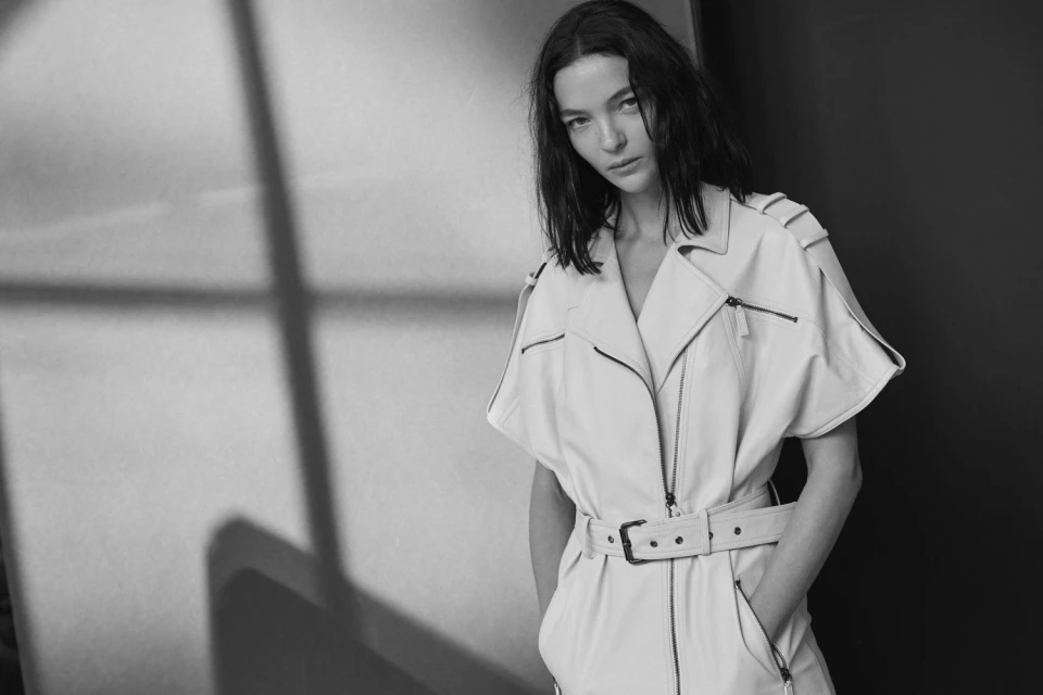 Monochrome portrait of a woman wearing a belted jumpsuit, standing in front of a wall with window-shadows on it. Studio lighting.