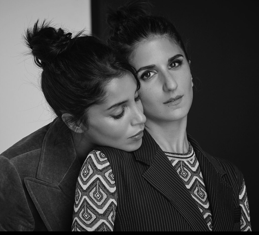 Black and white portrait of two women with their hair in buns wearing patterned tops and jackets in an indoor setting.