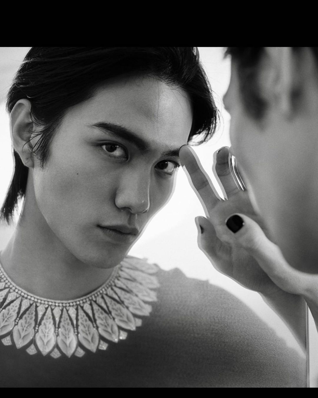Black and white portrait of a young adult male looking into a mirror, wearing an ornamental collar necklace, and touching the reflective surface.