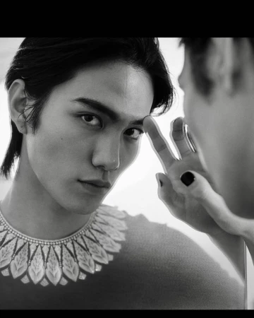 Black and white portrait of a young adult male looking into a mirror, wearing an ornamental collar necklace, and touching the reflective surface.