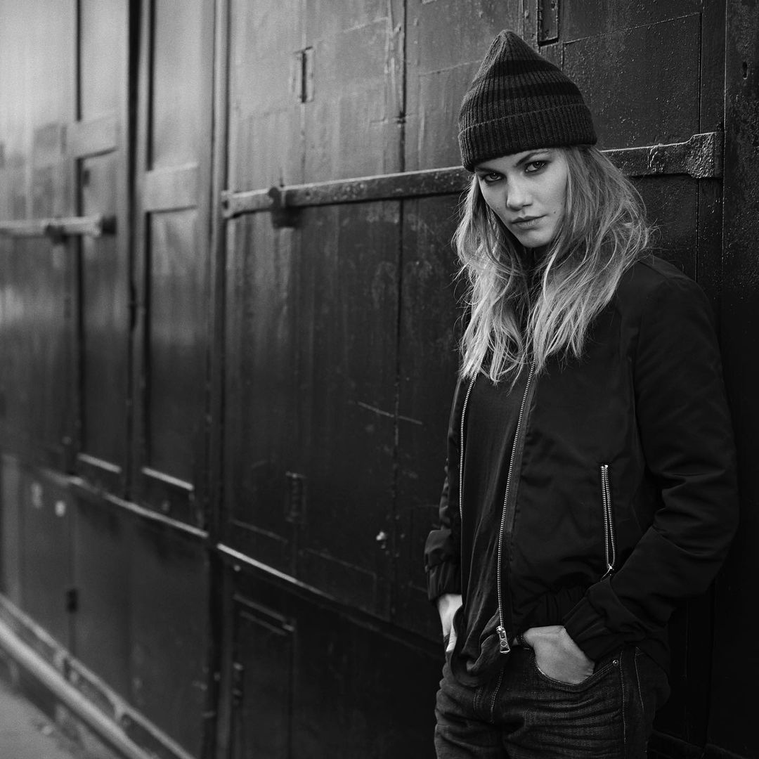 Black and white portrait of a woman wearing a ribbed beanie and a dark jacket leaning against a heavy door