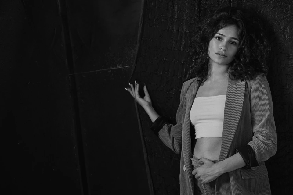 Portrait of a woman with curly hair wearing a corduroy suit in front of a dark textured background, creating a monochromatic aesthetic.
