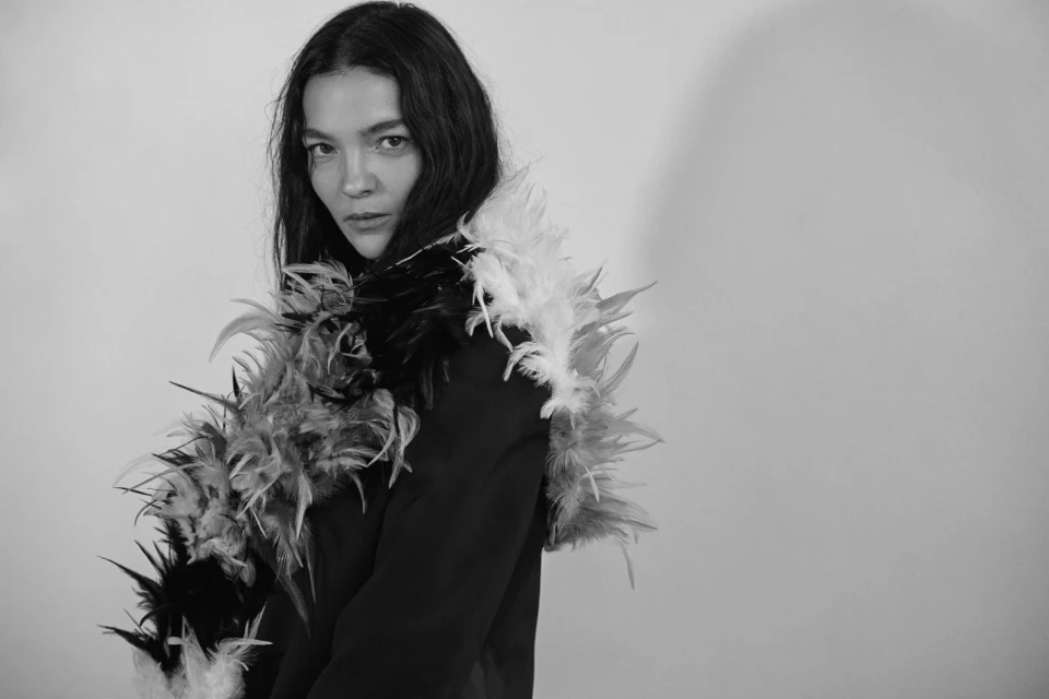 Monochrome portrait of a woman in a black jacket with a feathered collar, gazing at the camera against a stark white background.