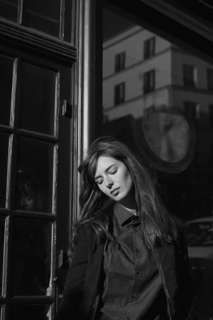 Black and white portrait of a woman with long hair leaning against a dark framed doorway with reflections of a building