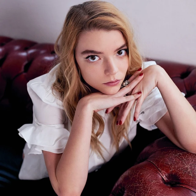 Blonde woman with red nails poses while resting her chin on her hands, wearing a white blouse and large ring, while sitting on a red couch.