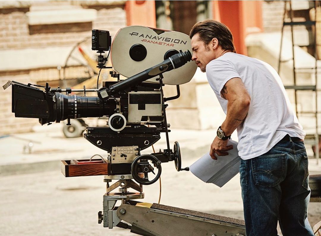 Man with a professional film camera on a set. He is looking into the viewfinder, dressed in a white shirt and blue jeans.