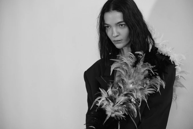 Monochrome portrait of a woman with dark wavy hair wearing a feathered jacket against a plain wall.