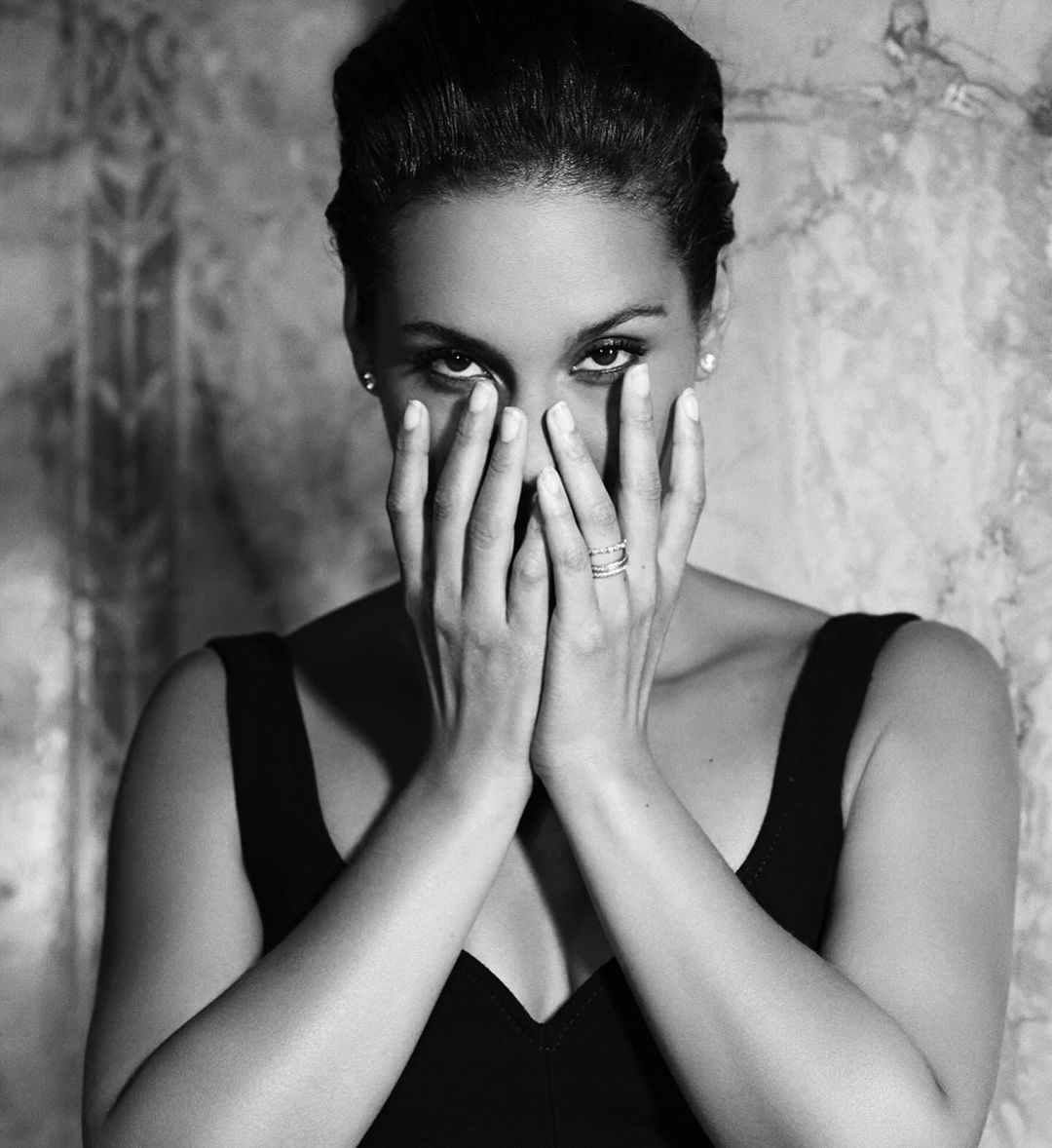 Black and white portrait of a woman with hands partially covering face. She wears a tank top and has dark hair pulled back.