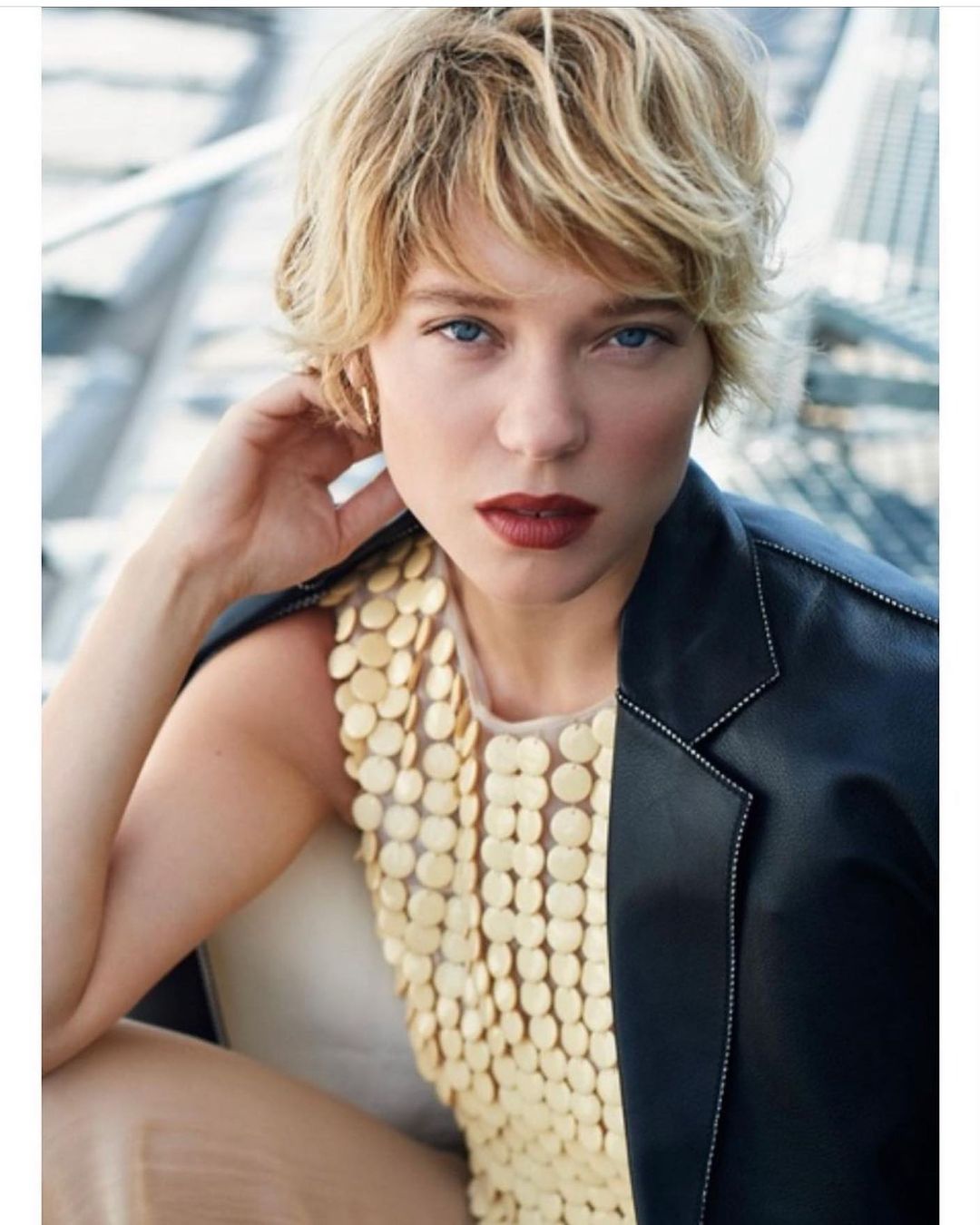 Blonde woman in leather jacket and gold sequin top posing indoors with shallow depth-of-field focus on the subject's face and shoulders.