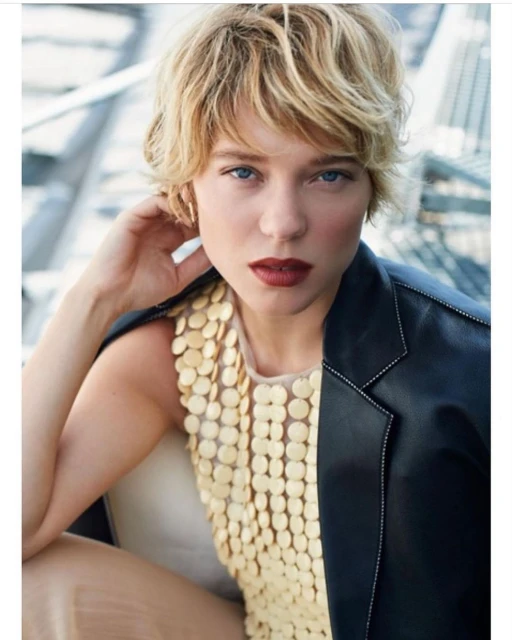 Blonde woman in leather jacket and gold sequin top posing indoors with shallow depth-of-field focus on the subject's face and shoulders.