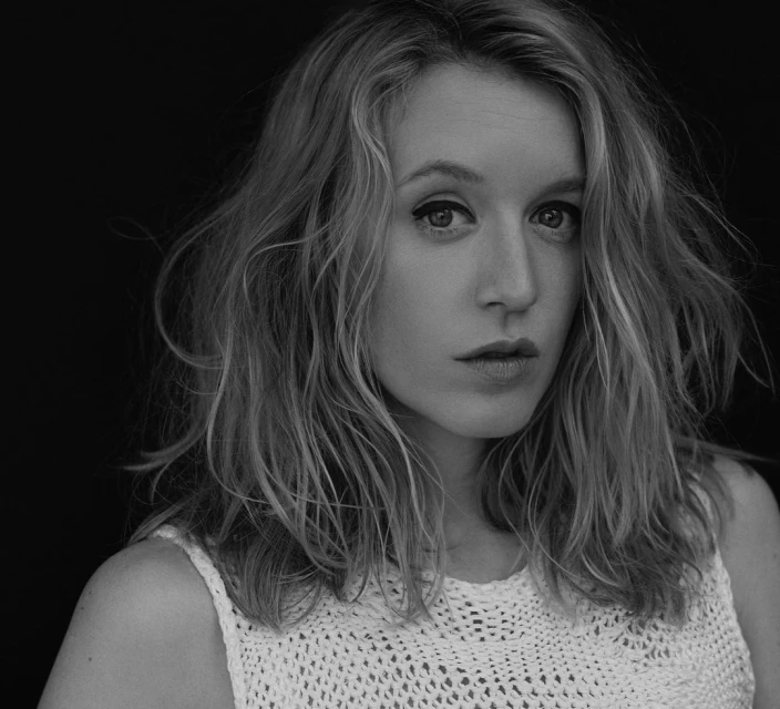 Black-and-white portrait of a blonde adult female looking at the camera, wearing a white crocheted top against a dark background.