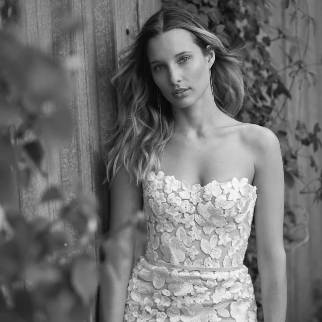 Black and white portrait of a young adult woman in a strapless dress with floral applique, standing near weathered wood and foliage.