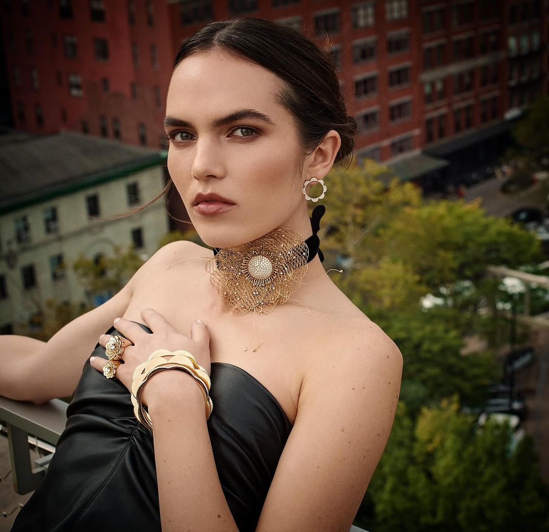Elegant woman poses in a black dress with gold jewelry against an urban backdrop of brick buildings and green trees.