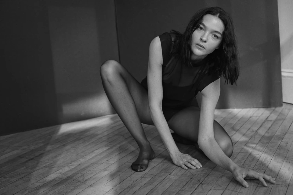 Monochrome portrait of a woman in a crouched pose wearing black tights and top against a dark gray wall and hardwood floor.