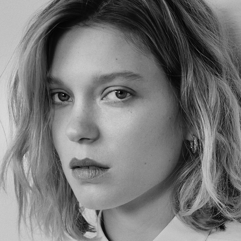 Close-up monochrome portrait of a woman with shoulder-length wavy hair and small hoop earrings looking at camera.