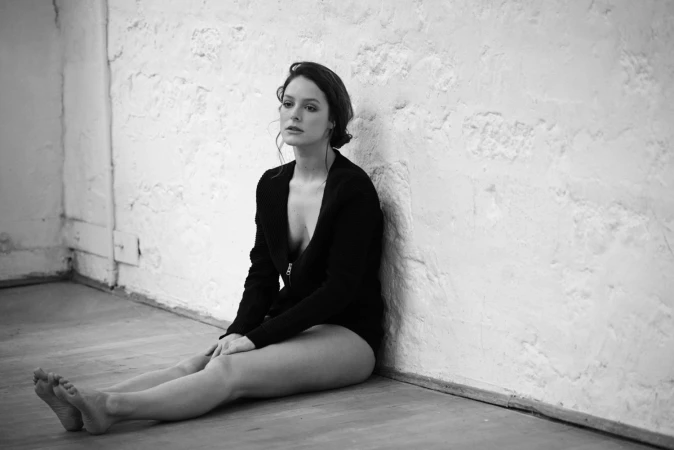 Woman in a black cardigan sitting barefoot on a wooden floor leaning against a textured white brick wall
