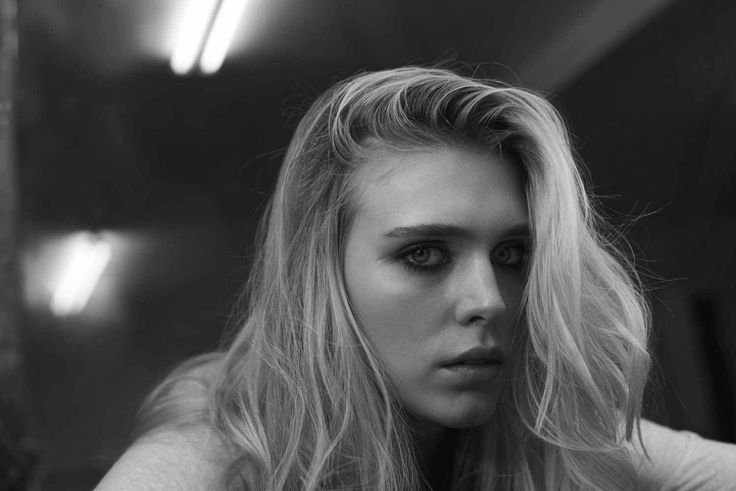 Close-up portrait of a young adult woman with long blonde hair, gazing directly at the camera in black and white with a diffused background.