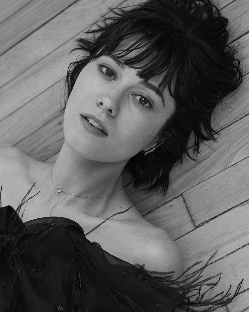Black-and-white portrait of a woman with dark hair lying on a wood floor wearing a black, feathered top, looking at the camera.