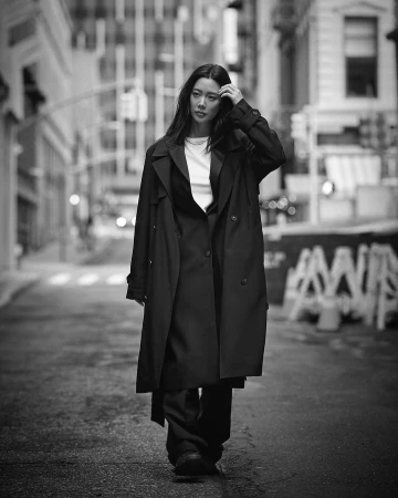 Asian woman in a trench coat walks down an urban street in black and white. City buildings and street barriers create a streetscape.