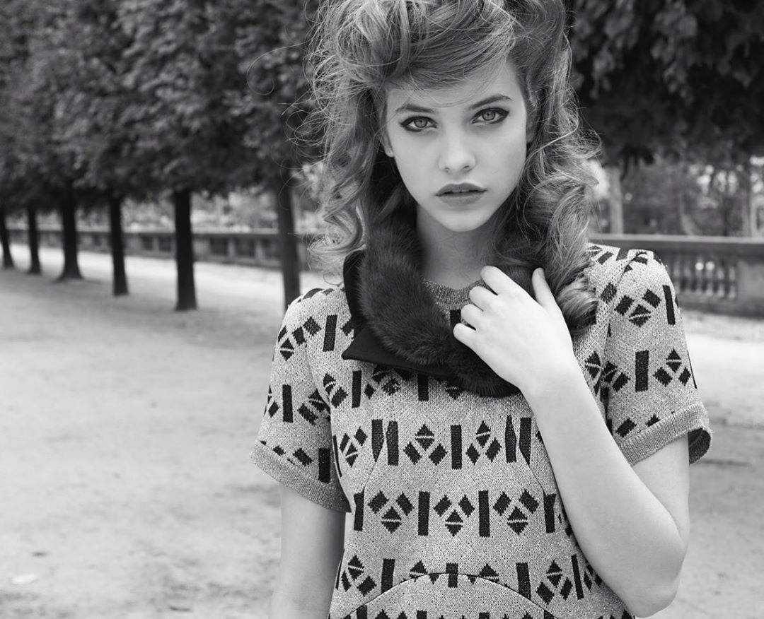 Black and white portrait of a young woman with wavy hair, wearing a patterned dress and fur collar, standing outdoors in a park setting.