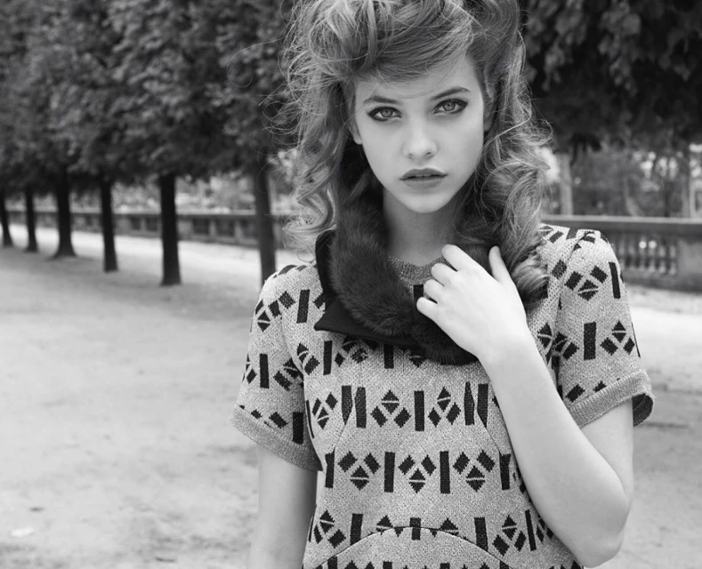 Black and white portrait of a young woman with wavy hair, wearing a patterned dress and fur collar, standing outdoors in a park setting.