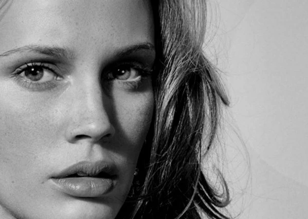 Close-up monochrome portrait of a woman with freckles and dark hair against a plain light background, photographed by Eric Guillemain.