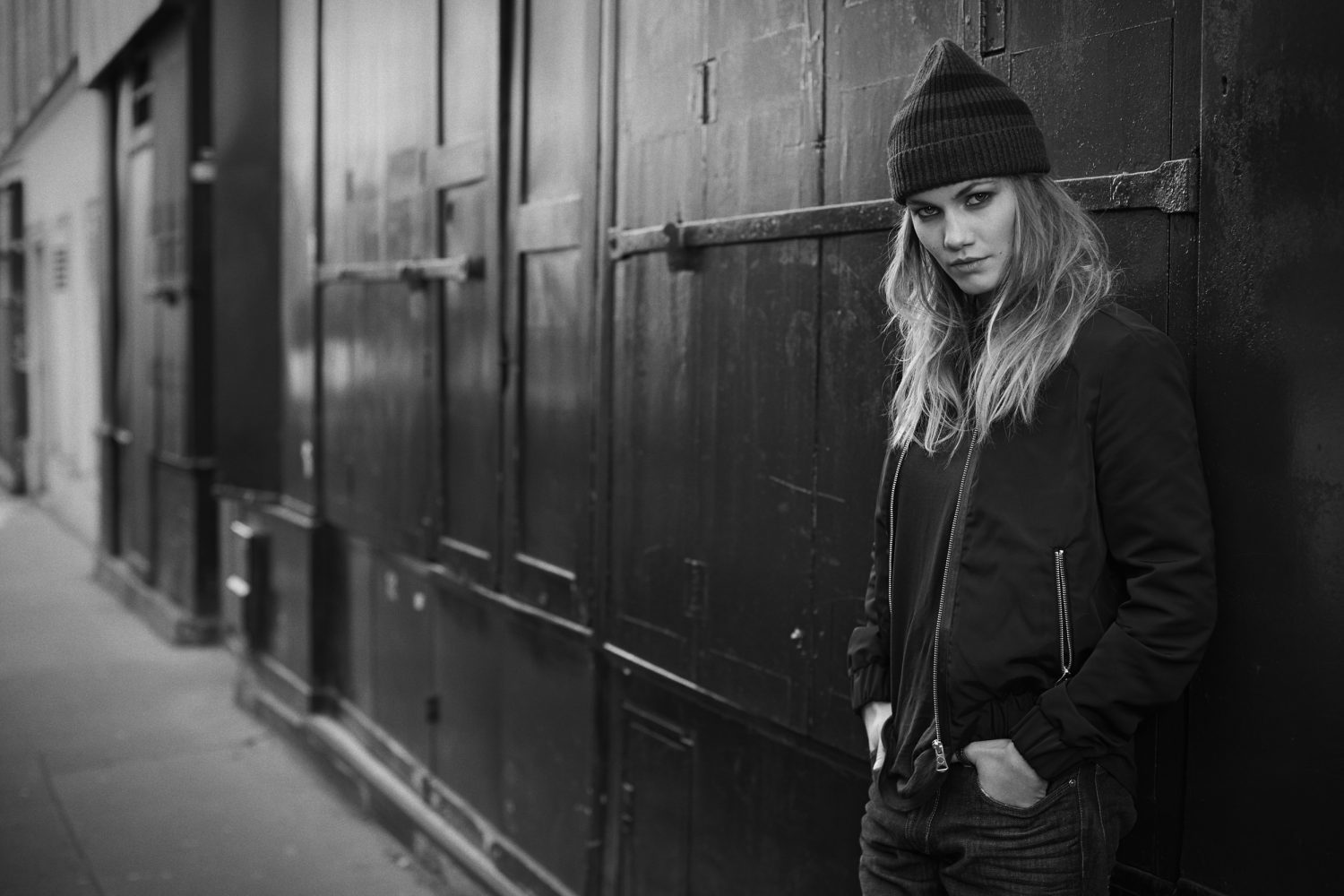 Black and white portrait of a young adult female in a beanie and jacket, leaning against a textured wall in an urban setting.