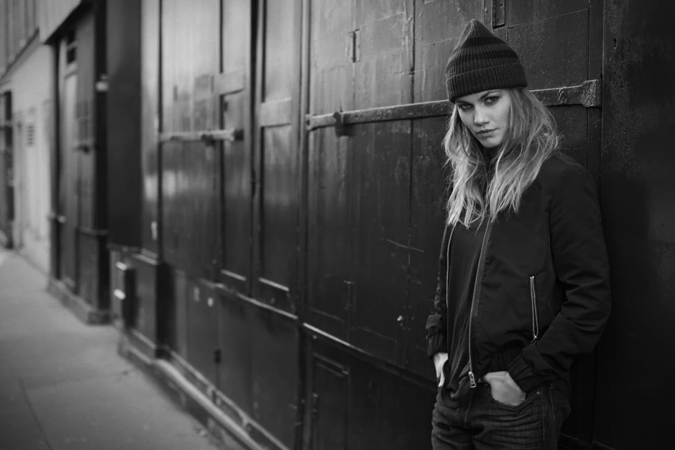 Black and white portrait of a young adult female in a beanie and jacket, leaning against a textured wall in an urban setting.
