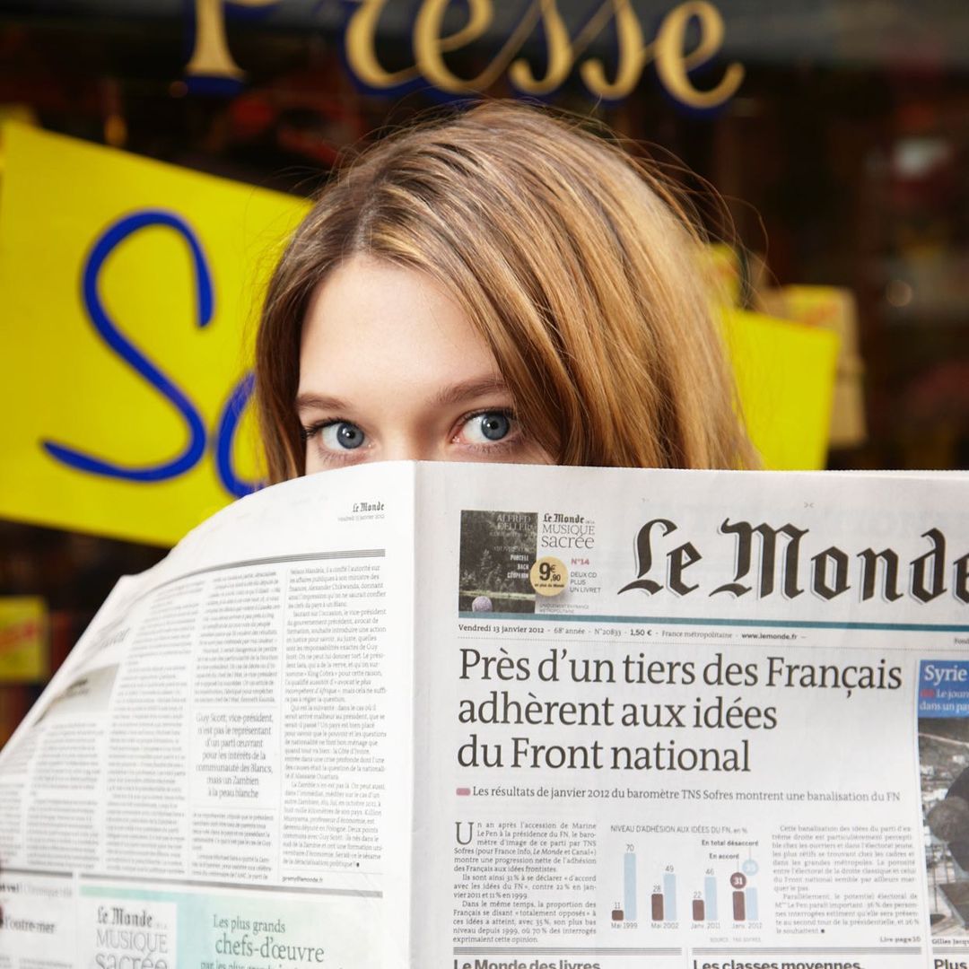 Close-up portrait of a woman holding a French newspaper hiding the lower half of her face by Eric Guillemain.