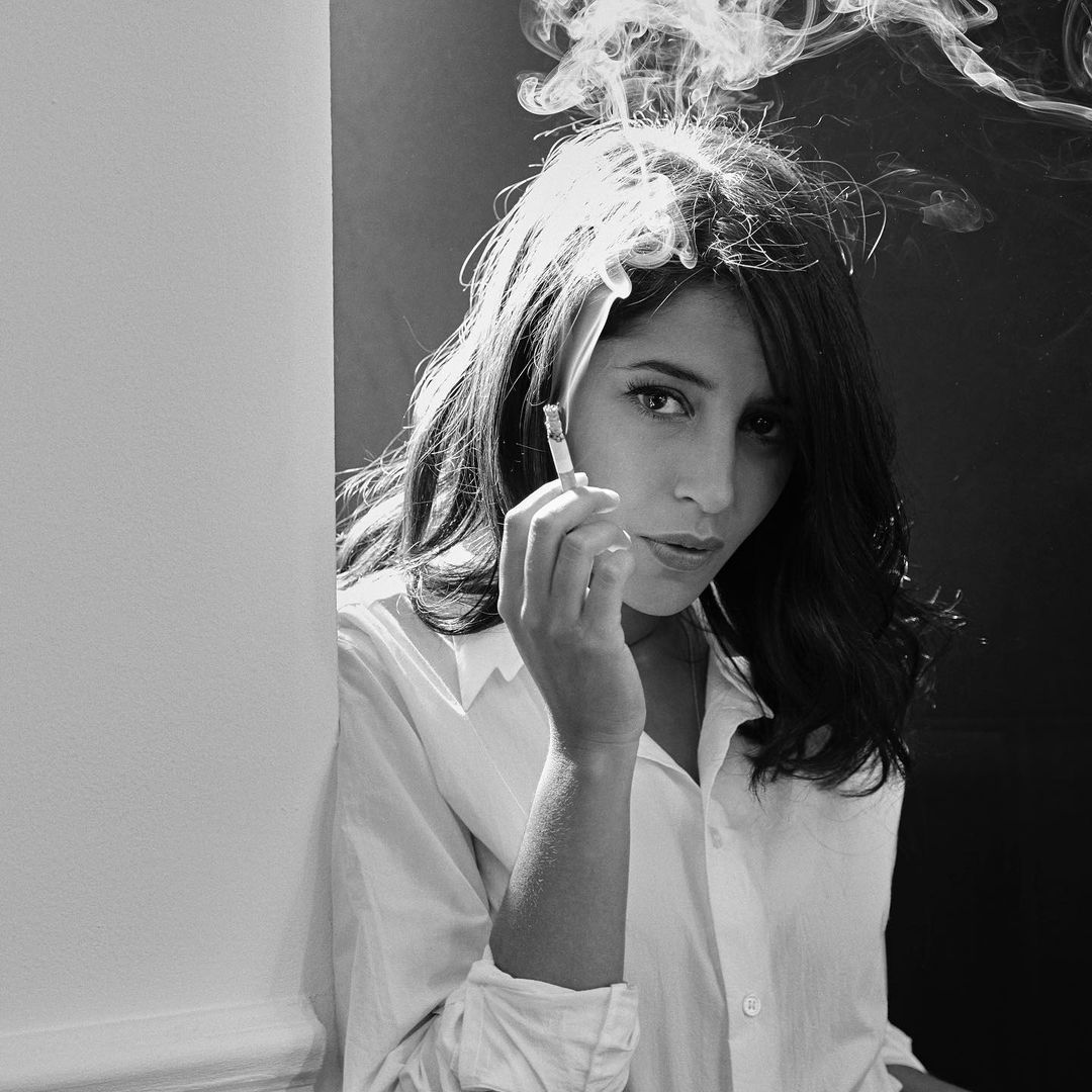 Portrait of a young woman with dark hair and a white shirt, smoking a cigarette with smoke curling up around her head in black and white.