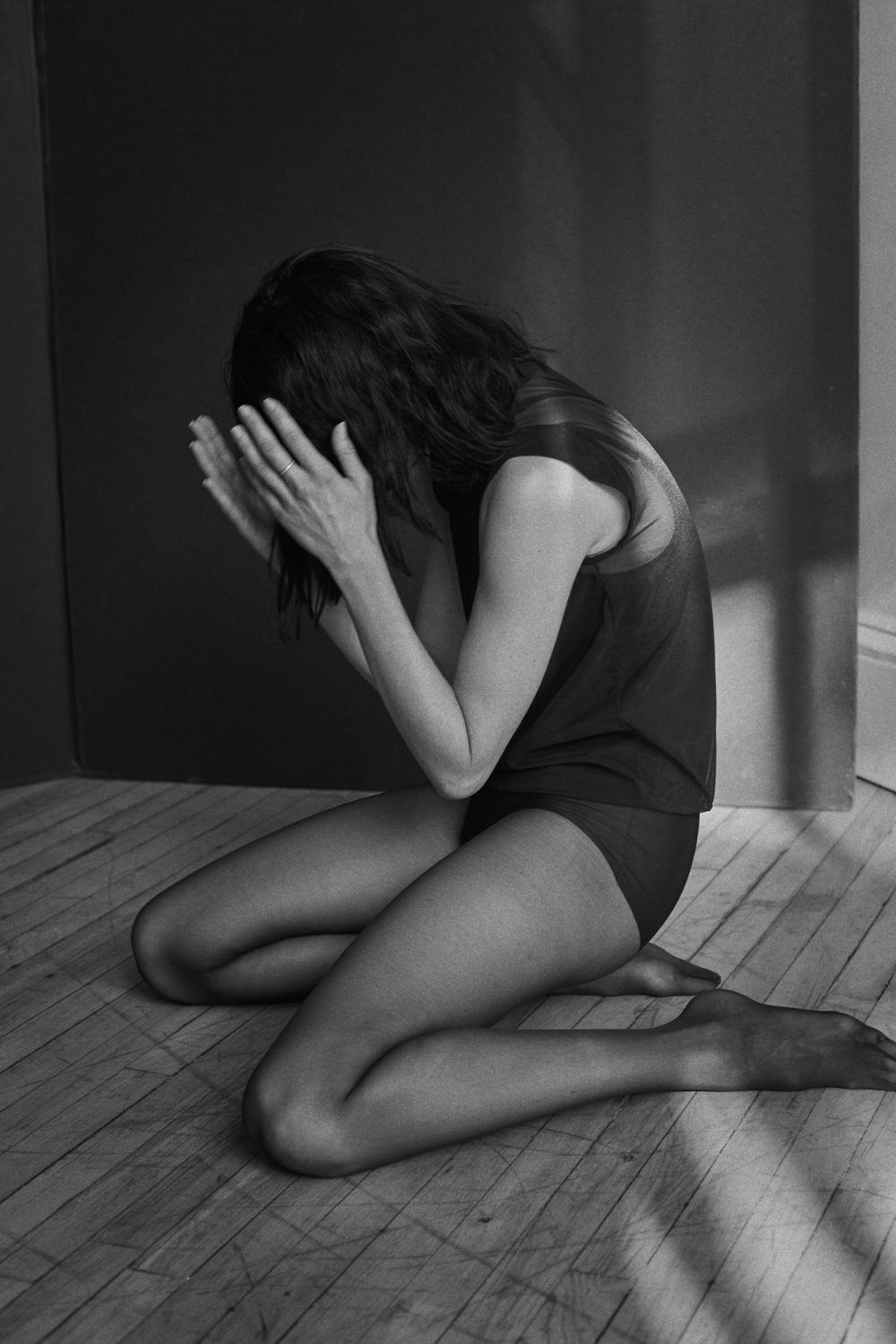 Black and white studio shot of a kneeling woman with head in her hands, wearing a dark top and tights on a wooden floor.