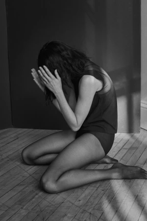 Black and white studio shot of a kneeling woman with head in her hands, wearing a dark top and tights on a wooden floor.