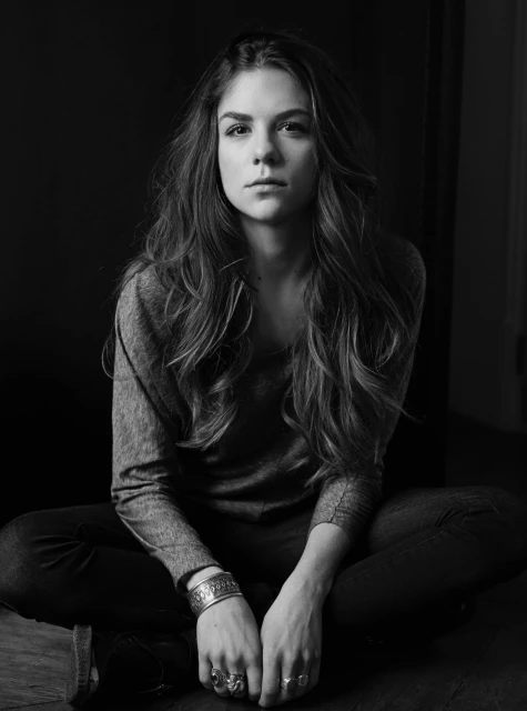 Black-and-white portrait of a young woman with long wavy hair sitting cross-legged, wearing a sweater, jeans, rings, and a bracelet.