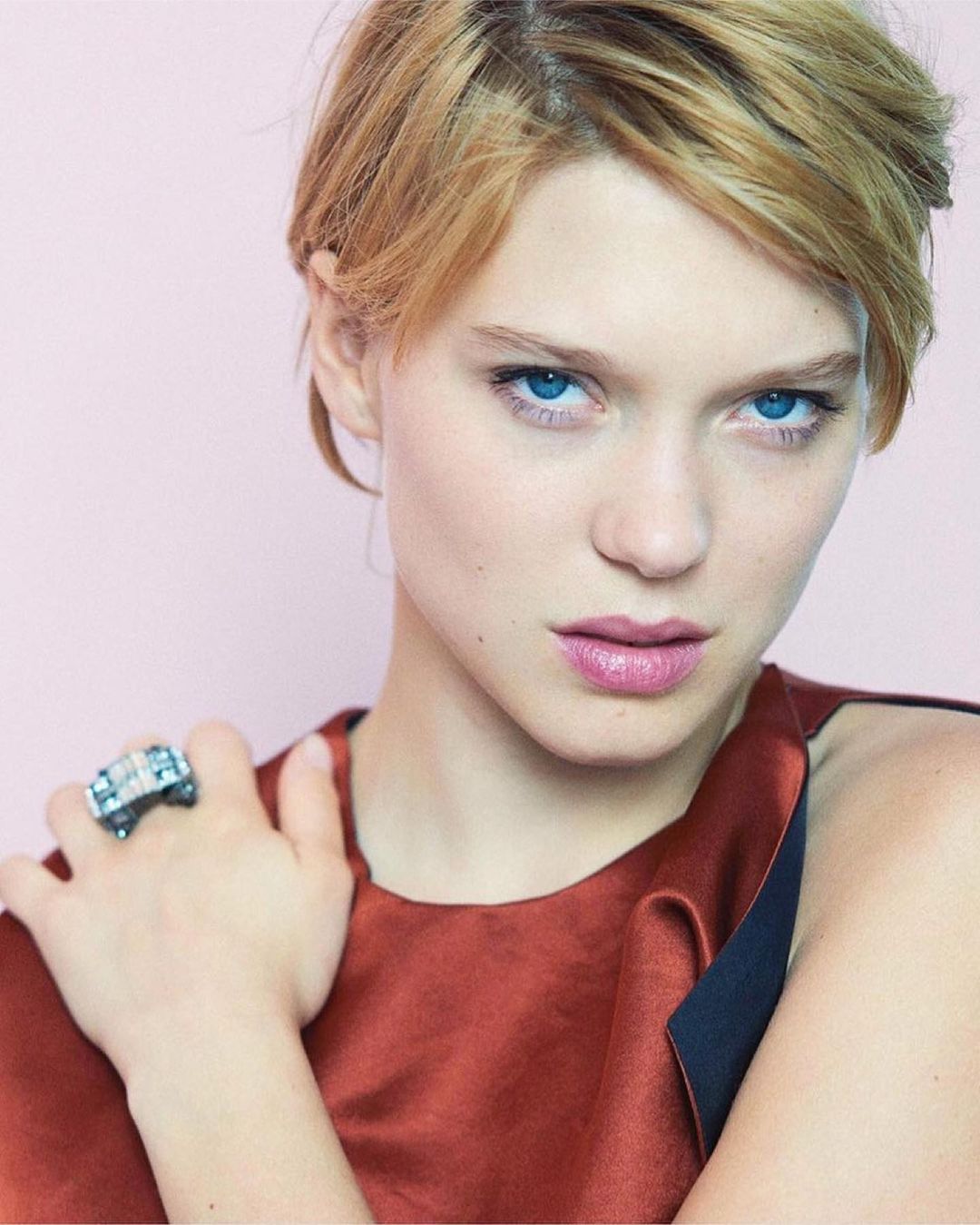 Close-up studio shot of a young adult woman with light skin and blonde hair, wearing a rust-colored top and a large, ornate ring.