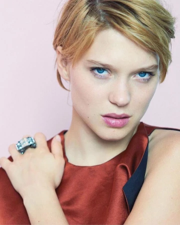 Close-up studio shot of a young adult woman with light skin and blonde hair, wearing a rust-colored top and a large, ornate ring.