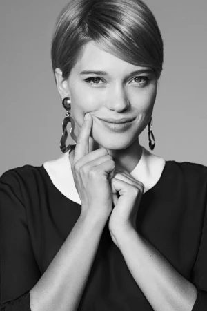 Portrait of Lea Seydoux with short hair smiling and pushing her cheek with a finger, wearing statement earrings.