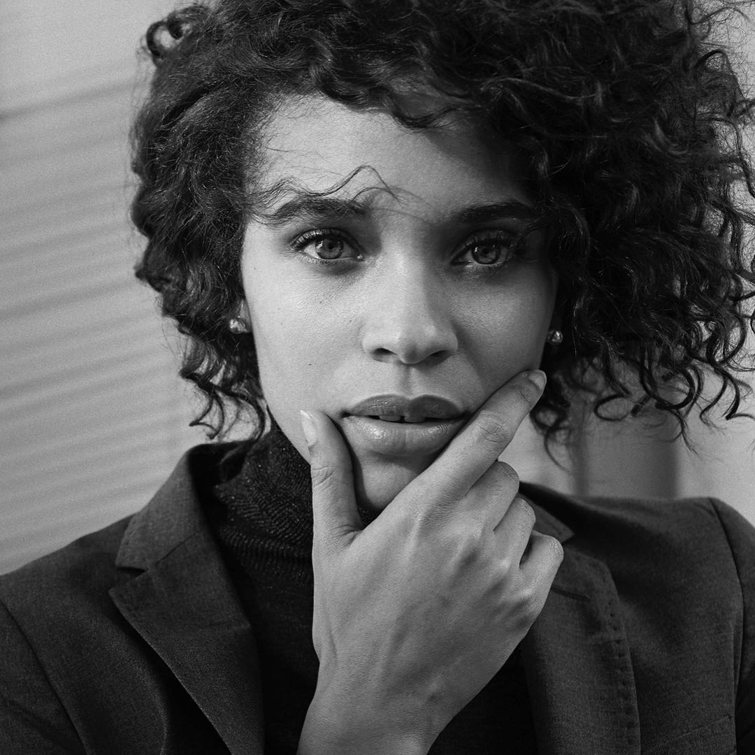 Black and white portrait of a woman with curly hair and a blazer looking at the camera with her hand on her chin.